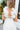 Upper back view of model wearing the Summer Romance Maxi Dress that has white fabric, maxi length, white lining, a ruffled hem, a plunge neckline, layered upper details, ruffle straps, and back corset ties