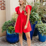 Full body front view of model wearing the Heart Breaker Dress In Red that has red fabric with a lining, a mini-length hem, a baby doll style, a v neckline, elastic straps with ruffles, and an open back with a bow detail