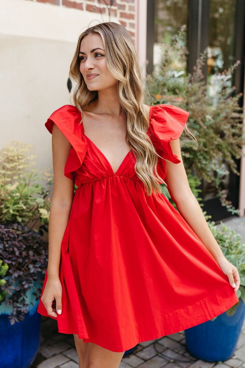 Front view of model wearing the Heart Breaker Dress In Red that has red fabric with a lining, a mini-length hem, a baby doll style, a v neckline, elastic straps with ruffles, and an open back with a bow detail