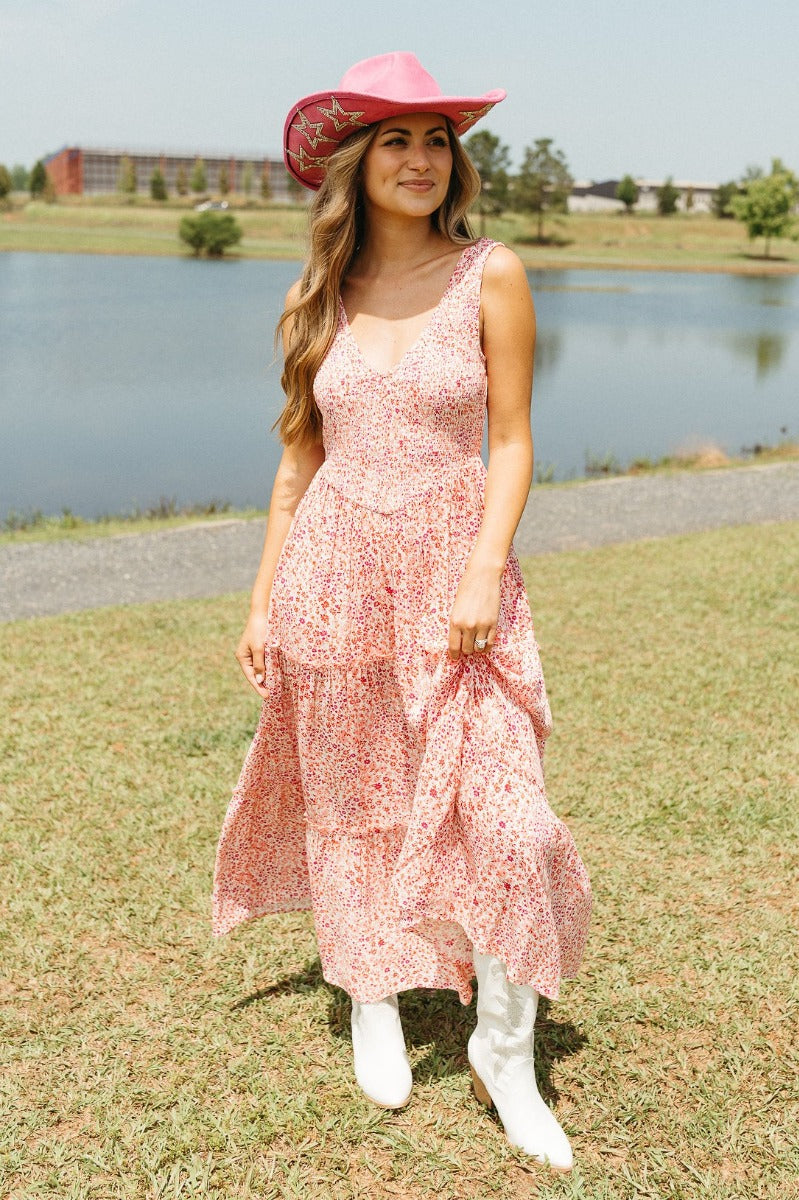 Full body front view of model wearing the Chasing Sunshine Floral Maxi Dress that has white fabric with a red and pink floral pattern, a two-tiered skirt, a smocked upper, a v-neck, sleeveless, and an open back with a tie closure
