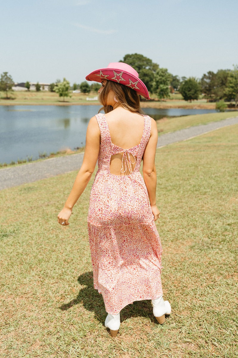 Full body back  view of model wearing the Chasing Sunshine Floral Maxi Dress that has white fabric with a red and pink floral pattern, a two-tiered skirt, a smocked upper, a v-neck, sleeveless , and an open back with a tie closure