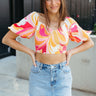 Front view of model wearing the Full of Happiness Top which features orange, fuchsia, pink, yellow and white fabric with a swirl pattern design, a cropped waist, a smocked body, a sweetheart neckline, and 3/4 sheer bubble sleeves with elastic wrists.