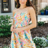 Front view of model wearing the Dream Of Vacation Dress which features a blue, teal, green, white, orange, pink and yellow floral print, mini length, white lining, a two-tiered body, a smocked upper, a strapless neckline with lettuce trim, and ties on the