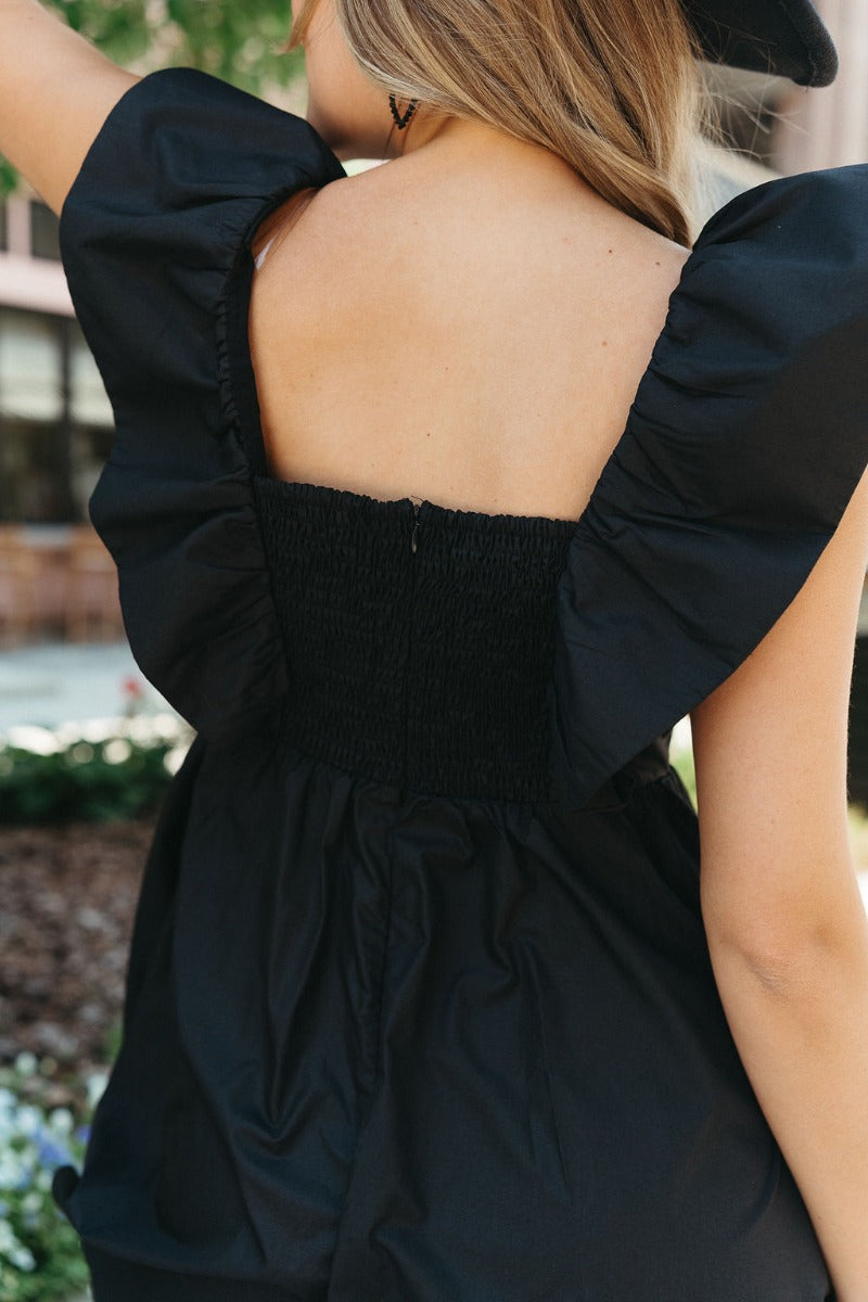 Close up back view of model wearing the Total Knockout Dress which features black fabric, mini length, a v-neckline, side pockets, pleated upper details, dramatic ruffle straps, black lining, a smocked upper and a monochromatic back zipper with a hook clo
