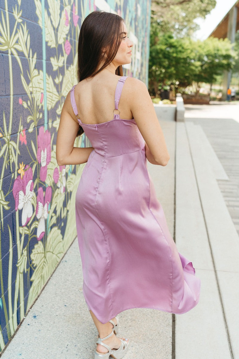 Back view of model wearing the Blaire Satin Midi Dress which features lavender satin fabric, midi length, a slit on the side, exterior boning and mesh details, pleated upper fabric, a sweetheart neckline, adjustable straps, and a monochromatic back zipper