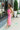 Full body side view of model wearing the One Step Ahead Midi Dress which features pink fabric, upper corset boning details, pink lining, midi length, side slits, a cowl neckline, adjustable straps, and a monochromatic back zipper with a hook closure.