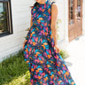 Full body front view of model wearing the Laguna Floral Maxi Dress that has a navy, orange, pink, blue, purple and green geometric/floral print, a tiered skirt, a smocked top, and a high neck with lettuce trim.