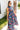 Full body side view of model wearing the Laguna Floral Maxi Dress that has a navy, orange, pink, blue, purple and green geometric/floral print, a tiered skirt, a smocked top, and a high neck with lettuce trim.