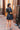 Back view of model wearing the Dream Come True Bubble Dress that has black fabric, mini length, a bubble hem, black lining, an elastic waist, a surplice neck and short puff sleeves.
