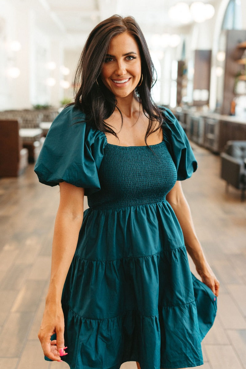 Front view of model wearing the Falling Fast Dress in Hunter Green which features teal fabric, mini length, two tiered baby doll style, teal lining, smocked chest, square neckline and short puff sleeves.