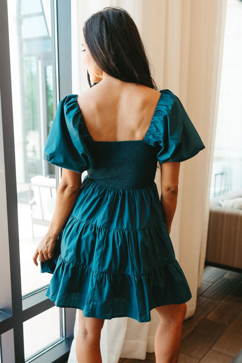 Back view of model wearing the Falling Fast Dress in Hunter Green which features teal fabric, mini length, two tiered baby doll style, teal lining, smocked chest, square neckline and short puff sleeves.