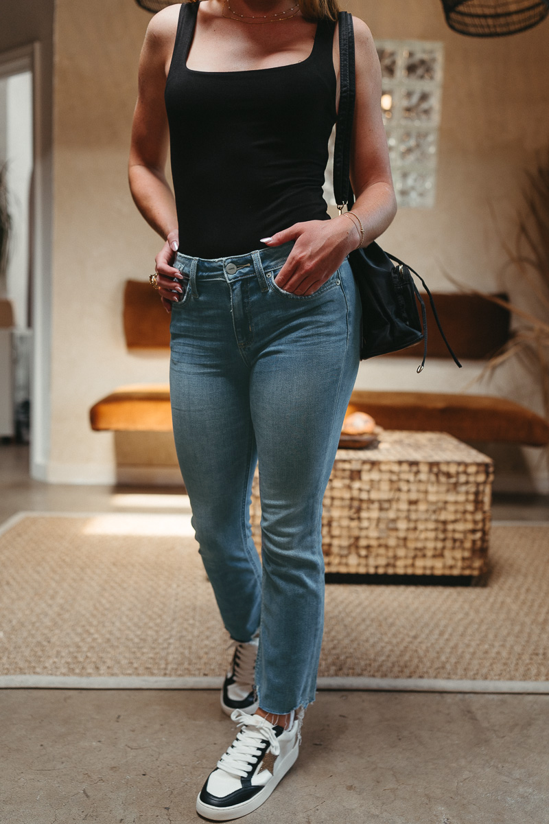 Front view of model wearing theThe Ceros: Meet Me Downtown Jeans which features medium blue denim wash, front zipper with button closure, mid-rise, two front pockets, two back pockets, belt loops and cropped flare with distressed hem.