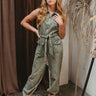 Front view of model wearing the Road Trip Denim Jumpsuit that has olive acid wash denim fabric, front pockets, belt loops with tie around the waist, straight leg pants, a front zipper, a collar, and sleeveless.