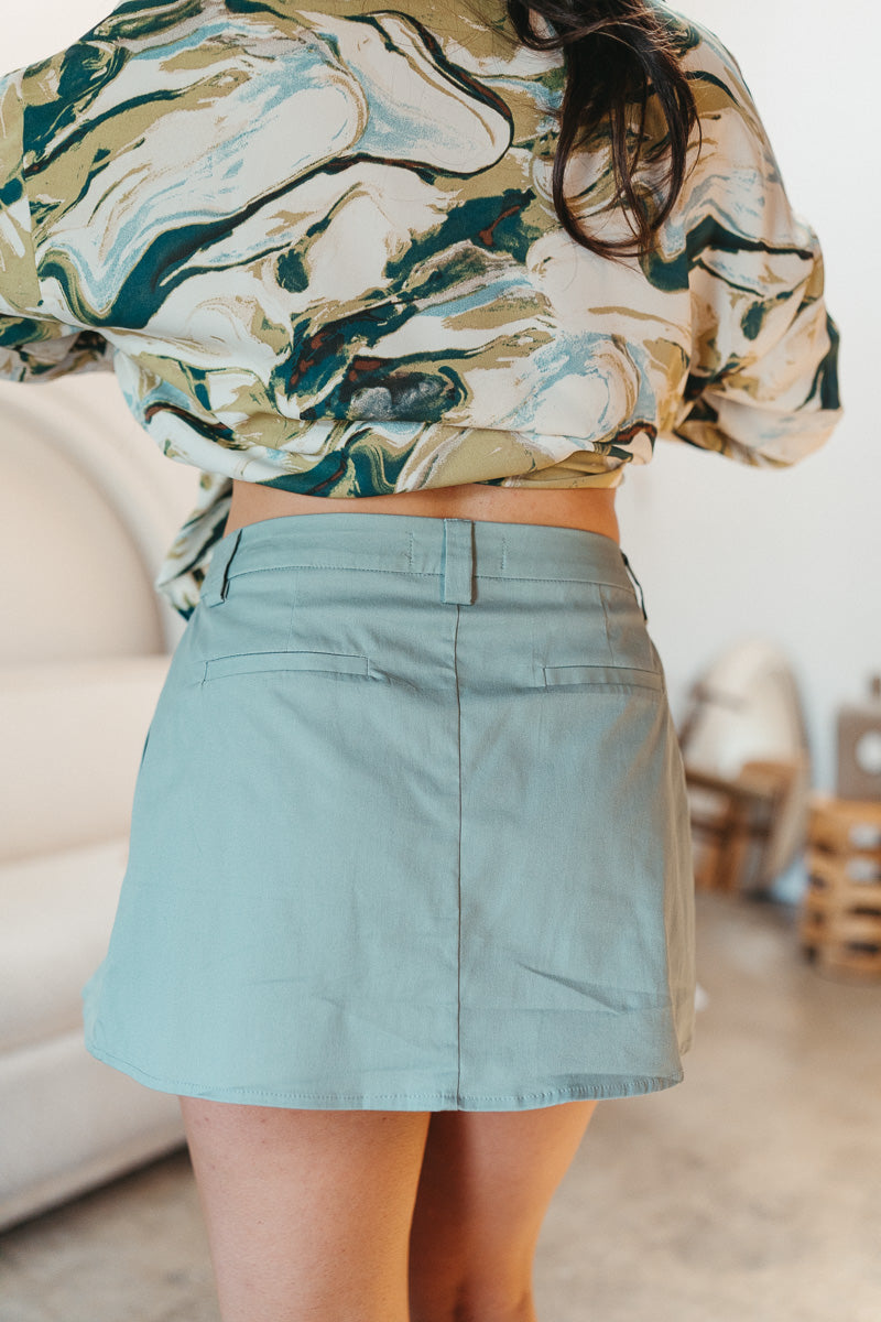 Back view of model wearing the Santa Monica Pleated Skirt that has sage fabric, pleated details, a front zipper with a hook closure, belt loops, and back pockets