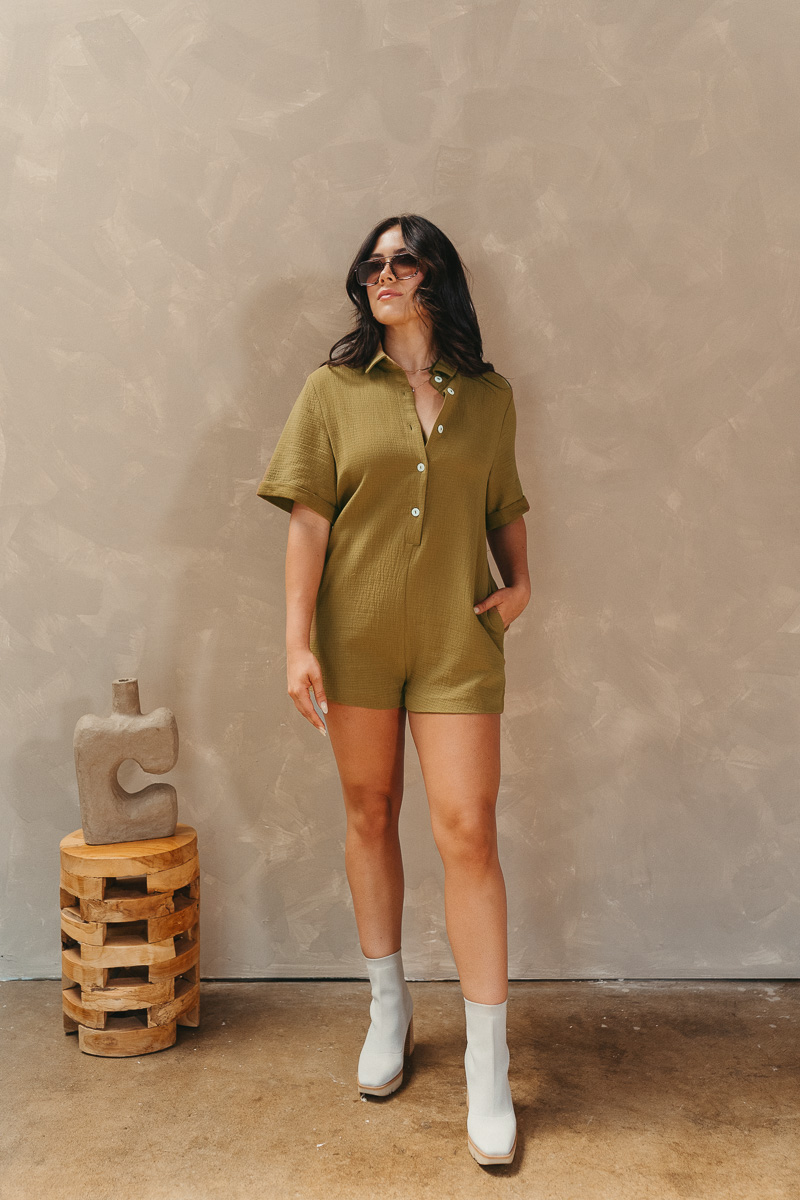 Full body view of model wearing the Western Winds Romper that has olive gauze fabric, pearlescent buttons, a collared neckline, two front pockets, and short cuffed sleeves.
