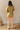 Full body back view of model wearing the Western Winds Romper that has olive gauze fabric, pearlescent buttons, a collared neckline, two front pockets, and short cuffed sleeves.