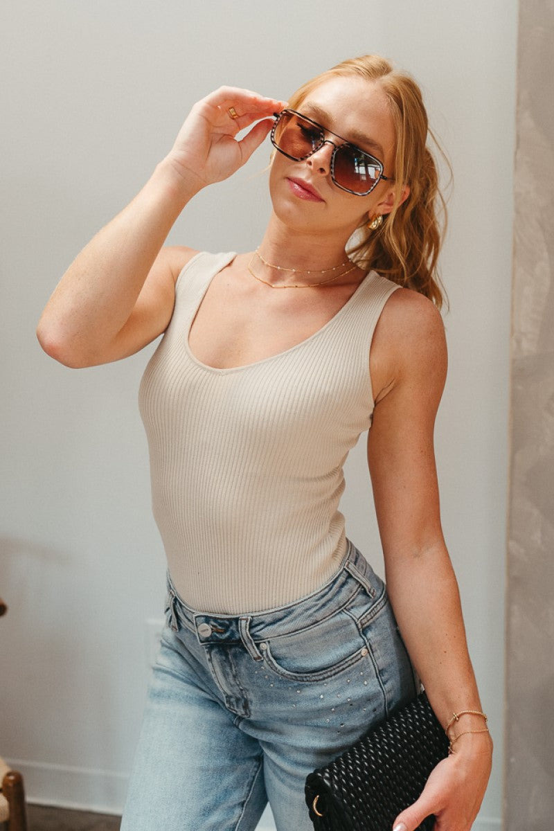 Front view of model wearing the Let's Go Ribbed Bodysuit that has oatmeal ribbed knit fabric, a scooped neckline, a sleeveless body, and a thong bottom with adjustable snap closures