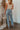 Back view of model wearing the Ceros: Aurora Embellished Jeans that have light blue denim wash fabric, rhinestone details, front zippe, belt loops, pockets, brown stitching, and straight legs.