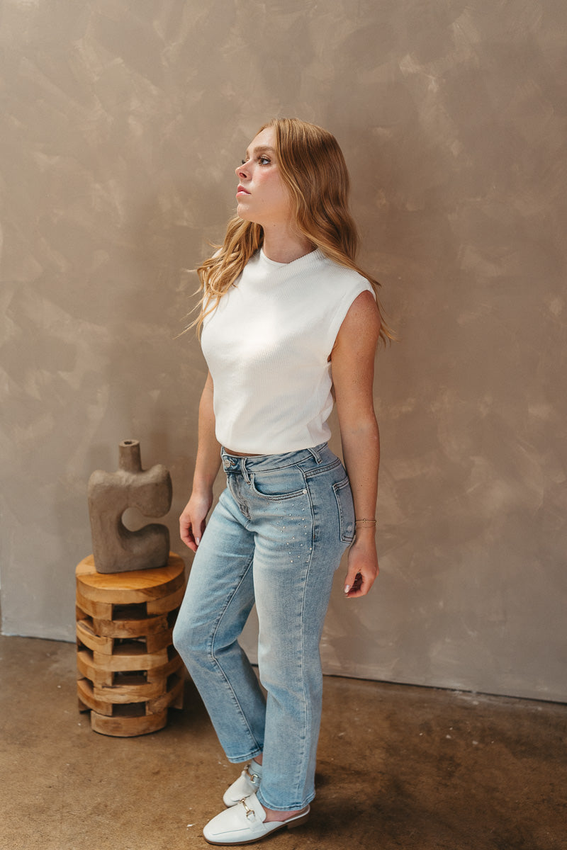 Full body side view of model wearing the Keep Dreaming Top which features white knit fabric, a high neckline, a thick hem, and a sleeveless design with shoulder pads.