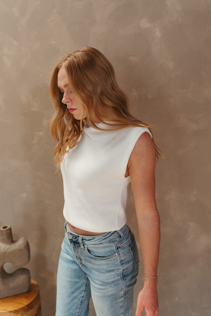 Side view of model wearing the Keep Dreaming Top which features white knit fabric, a high neckline, a thick hem, and a sleeveless design with shoulder pads.