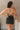 Back view of model wearing the Spin Me Around Romper which features washed black denim, two front pockets, a sweetheart neckline, adjustable straps, an open back with corset ties, and a back zipper.