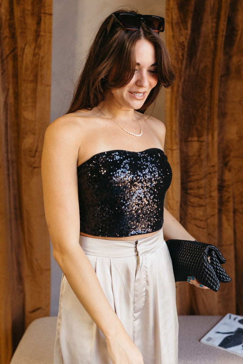 Frontal side view of model wearing the Maya Black Sequin Tube Top that has black sequin fabric, a cropped waist, a strapless sweetheart neckline, and a back silver zipper closure.<