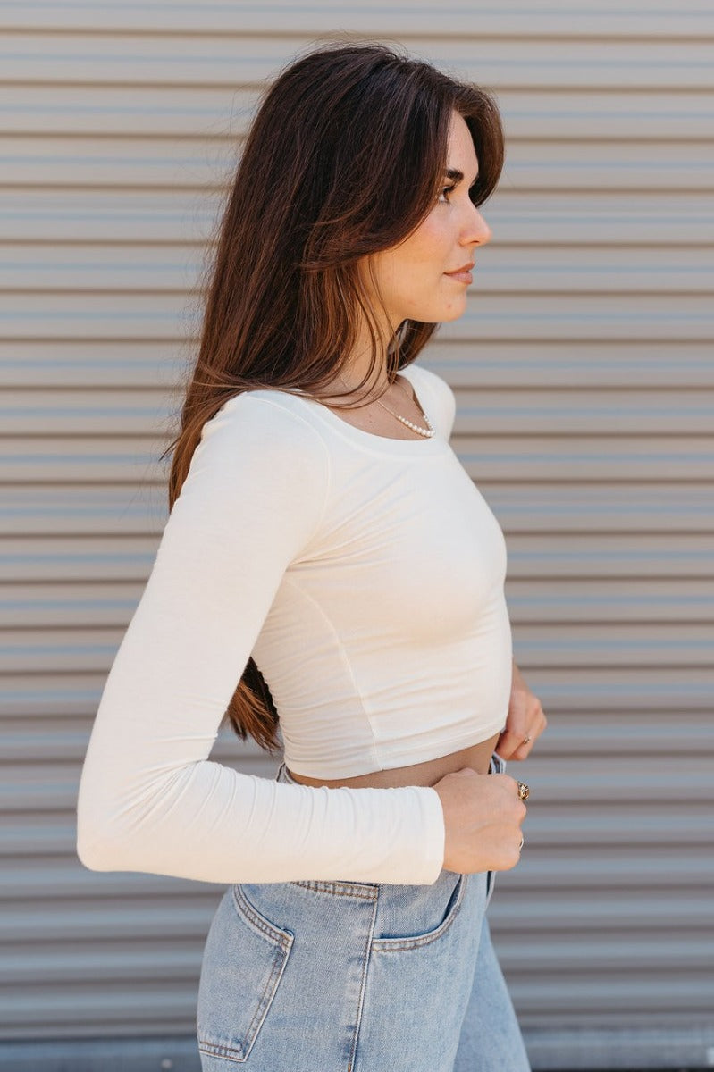 Side view of model wearing the The On Your Mind Top in Cream that has cream knit fabric, a cropped waist, a round neckline and long sleeves.
