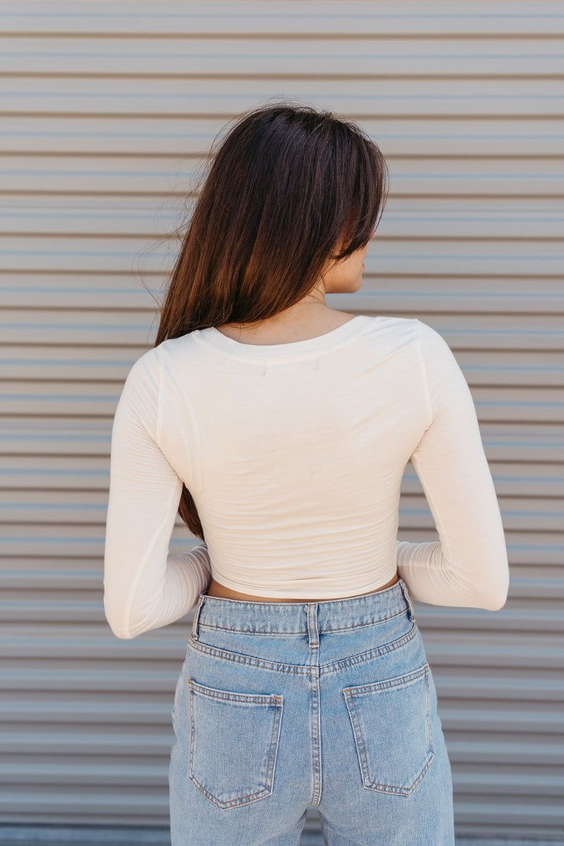 Back view of model wearing the The On Your Mind Top in Cream that has cream knit fabric, a cropped waist, a round neckline and long sleeves.
