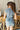 Back view of model wearing the Dallas Belted Zip-Front Denim Romper that has blue denim, pockets, a front zipper, a collar, an adjustable belt with a western silver buckle, and short cap sleeves