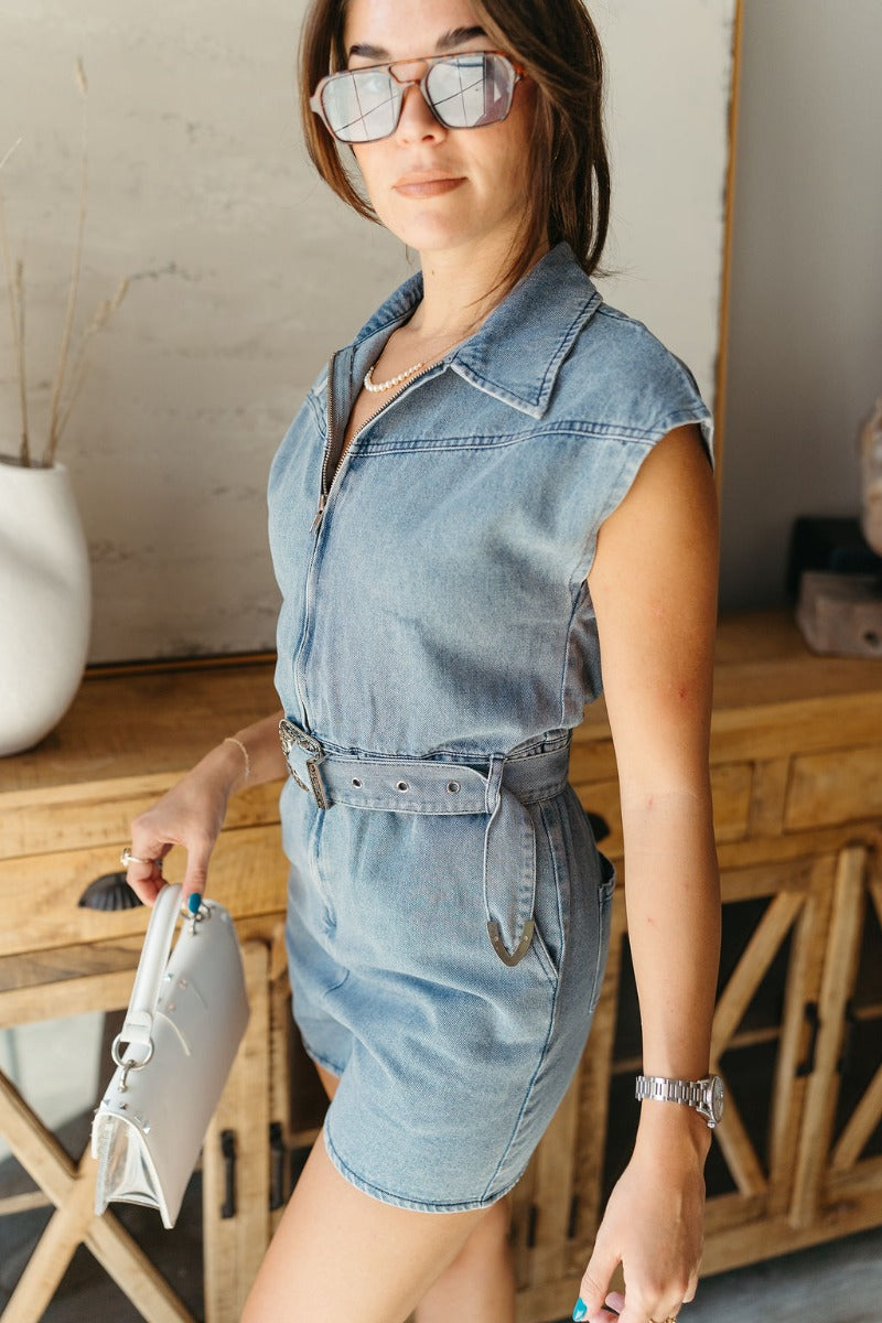 Side view of model wearing the Dallas Belted Zip-Front Denim Romper that has blue denim, pockets, a front zipper, a collar, an adjustable belt with a western silver buckle, and short cap sleeves
