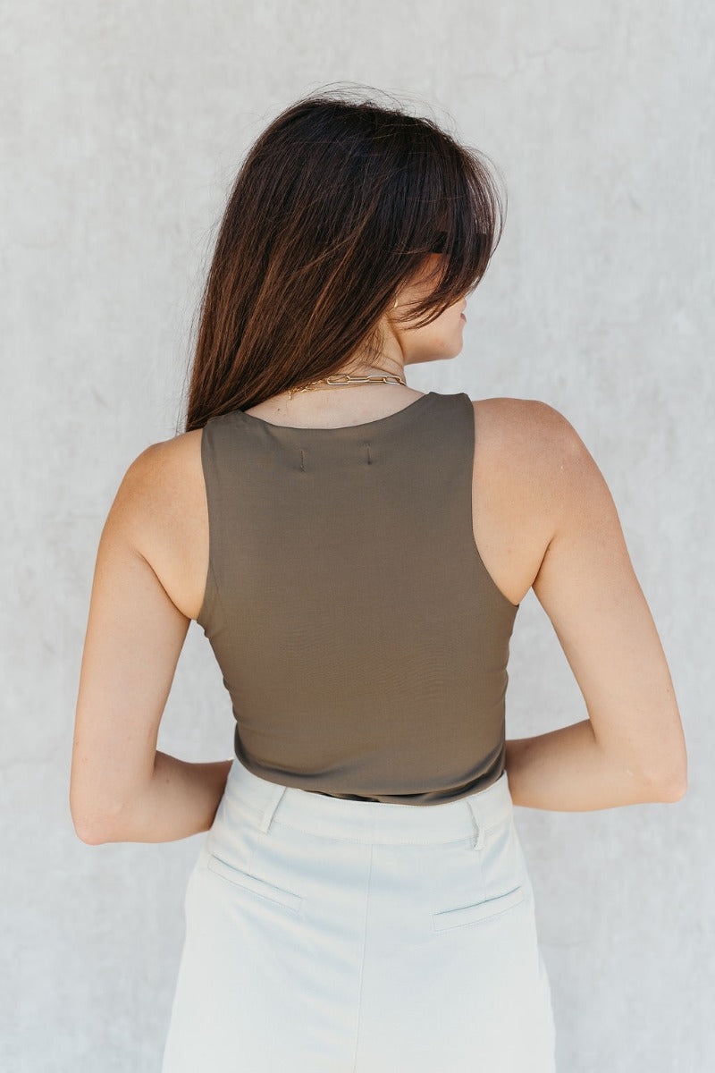 Back view of model wearing the Grace Olive Sleeveless Bodysuit which features dark olive knit fabric, a round neckline, a sleeveless design, and a thong bottom with snap closures.