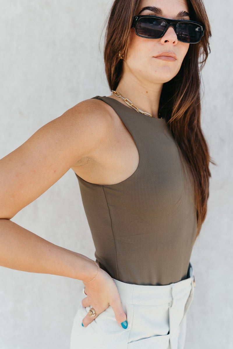 Side view of model wearing the Grace Olive Sleeveless Bodysuit which features dark olive knit fabric, a round neckline, a sleeveless design, and a thong bottom with snap closures.