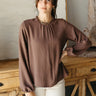 Front view of model wearing the Tillie Brown Long Sleeve Top that has lightweight brown fabric, a round neck with ruffle trim, a back key hole, and long sleeves with elastic wrists.