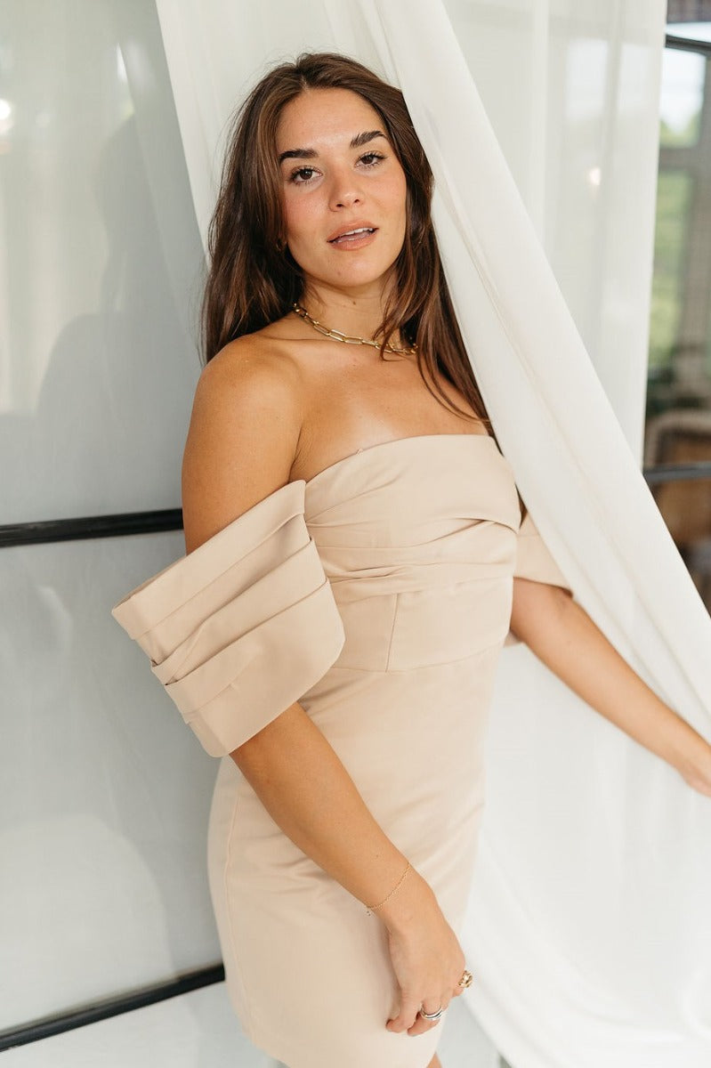 Side view of model wearing the Love Song Beige Off-The-Shoulder Mini Dress that has beige fabric, mini length, a pleated neckline, and off-the-shoulder pleated short sleeves.
