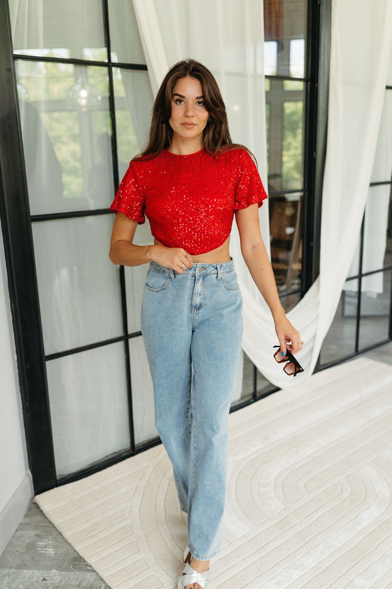 Full body front view of model wearing the Lennox Red Sequin Crop Top that has red sequin fabric, a cropped waist with an angled hem, a round neckline, short sleeves, and a silver back zipper closure.