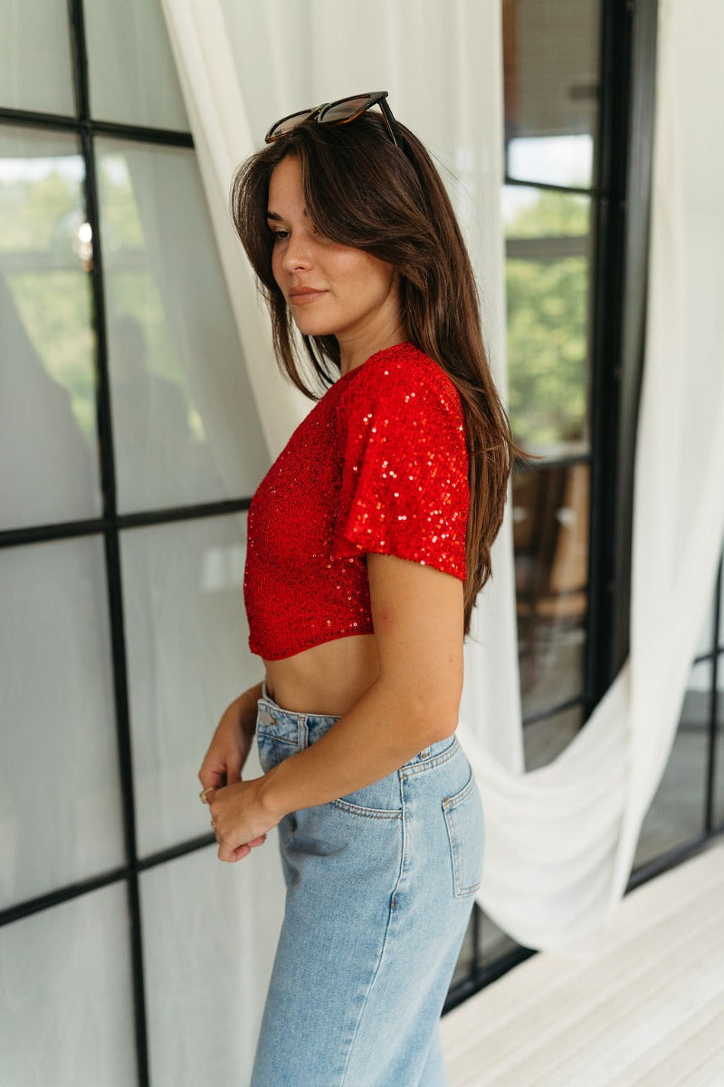 Side view of model wearing the Lennox Red Sequin Crop Top that has red sequin fabric, a cropped waist with an angled hem, a round neckline, short sleeves, and a silver back zipper closure.
