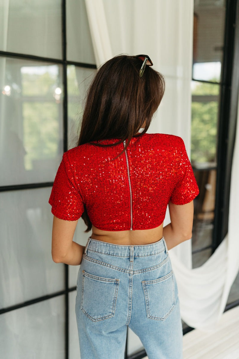 Back view of model wearing the Lennox Red Sequin Crop Top that has red sequin fabric, a cropped waist with an angled hem, a round neckline, short sleeves, and a silver back zipper closure.