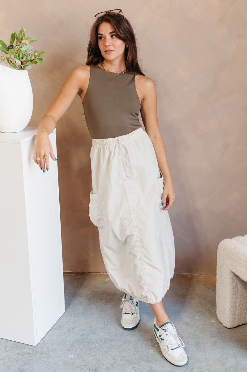 Full body front view of model wearing the Alina Cream Parachute Cargo Midi Skirt that has cream lightweight fabric, midi length, a back slit, two side pockets, an elastic drawstring waistband, and a drawstring hem.