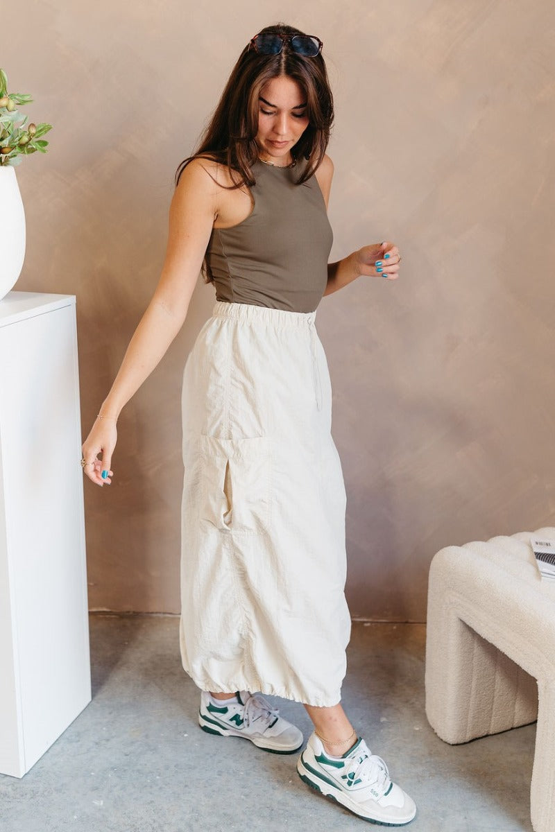 Full body side view of model wearing the Alina Cream Parachute Cargo Midi Skirt that has cream lightweight fabric, midi length, a back slit, two side pockets, an elastic drawstring waistband, and a drawstring hem.