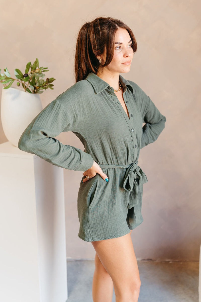 Side view of model wearing the Naomi Dark Green Long Sleeve Romper which features dark green knit fabric, two front pockets, monochromatic button up, collared neckline, tie around the waist and long sleeves with buttoned cuffs.