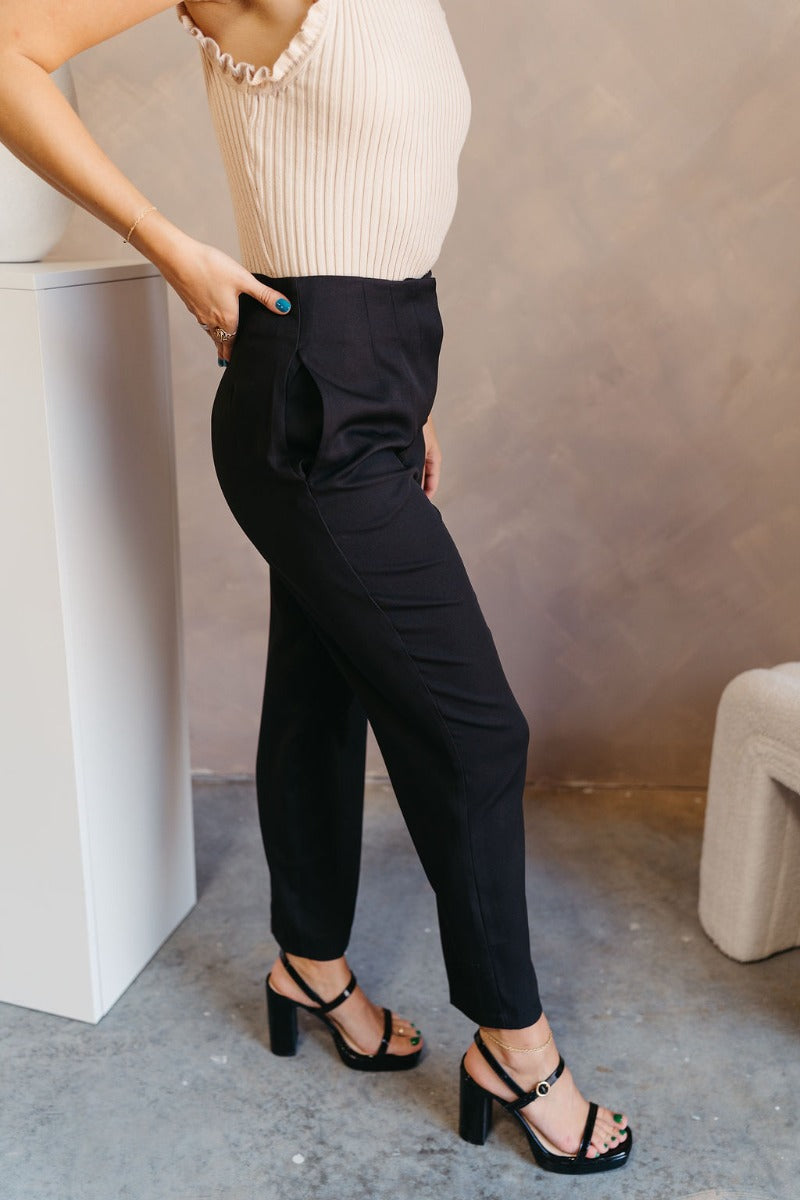 Side view of model wearing the Tessa Black Dress Pants which features black lightweight fabric, two front pockets, a front zipper with a hook closure, and tapered legs.