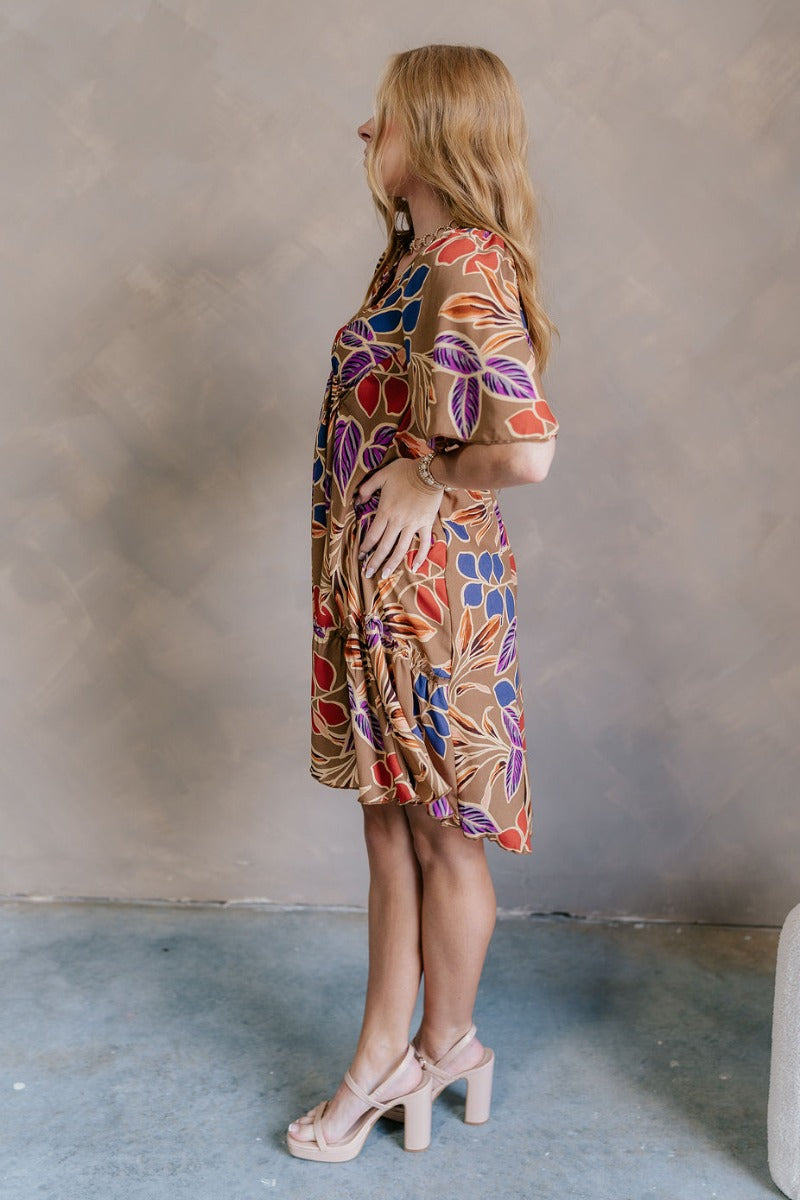 Side view of model wearing the Harper Multi Floral Mini Dress features light brown fabric with a blue, purple, rust and orange floral pattern, a ruffle hem skirt, a plunge neckline, a tie in the back, and short flare sleeves.