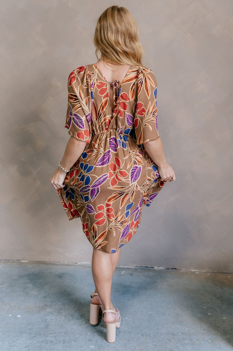 Back view of model wearing the Harper Multi Floral Mini Dress features light brown fabric with a blue, purple, rust and orange floral pattern, a ruffle hem skirt, a plunge neckline, a tie in the back, and short flare sleeves.