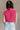 Back view of model wearing the Kelly Pink Cropped Turtleneck Sweater which features hot pink cable knit fabric, a cropped waist, a turtleneck neckline, and a sleeveless design.
