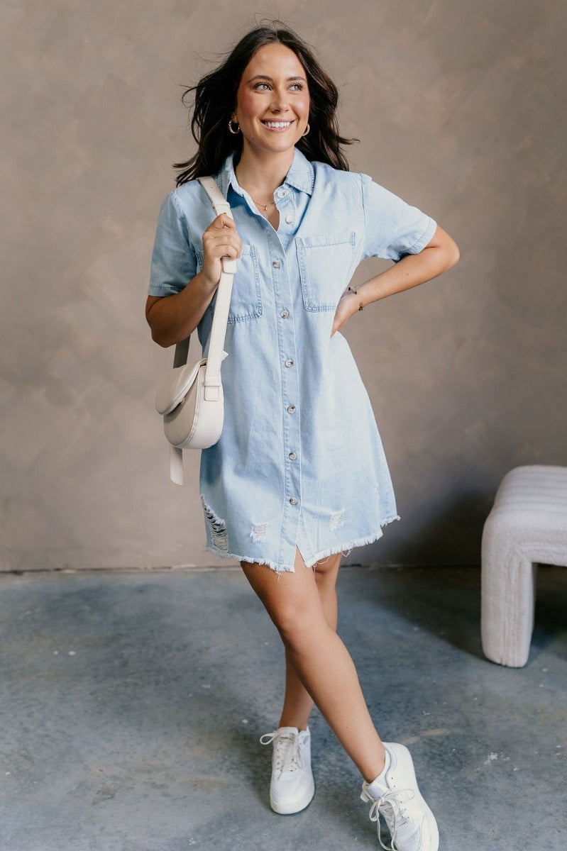 Full body front view of model wearing the Blakeley Distressed Chambray Mini Dress that has light blue chambray fabric, a frayed hem, distressed details, mini length, light tortoise buttons, pockets, a collared neck and short sleeves.