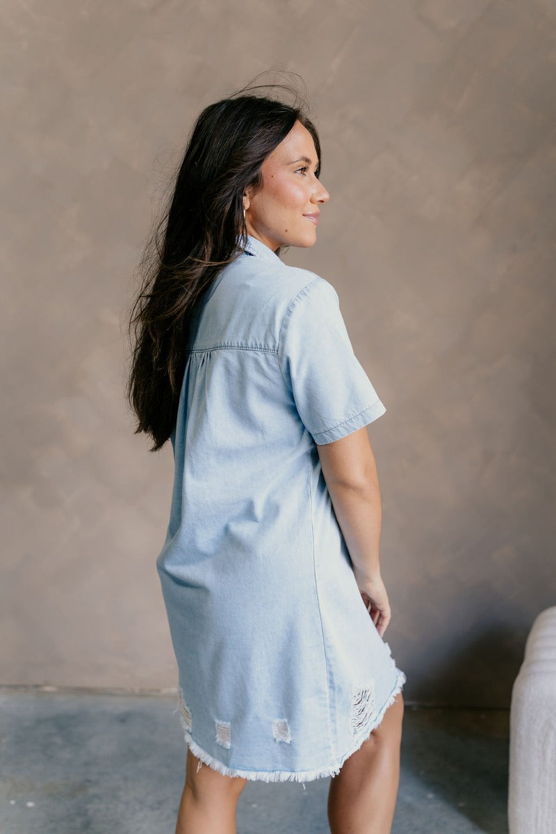 Side view of model wearing the Blakeley Distressed Chambray Mini Dress that has light blue chambray fabric, a frayed hem, distressed details, mini length, light tortoise buttons, pockets, a collared neck and short sleeves.