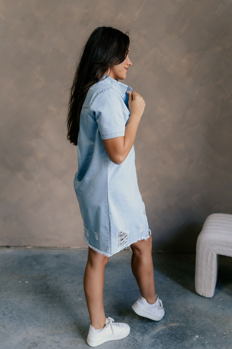 Full body side view of model wearing the Blakeley Distressed Chambray Mini Dress that has light blue chambray fabric, a frayed hem, distressed details, mini length, light tortoise buttons, pockets, a collared neck and short sleeves.