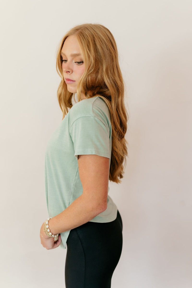 Side view of model wearing the Lillie Mint Green Basic Short Sleeve Top that has mint green knit fabric, a scooped hem, a round neckline and short sleeves.