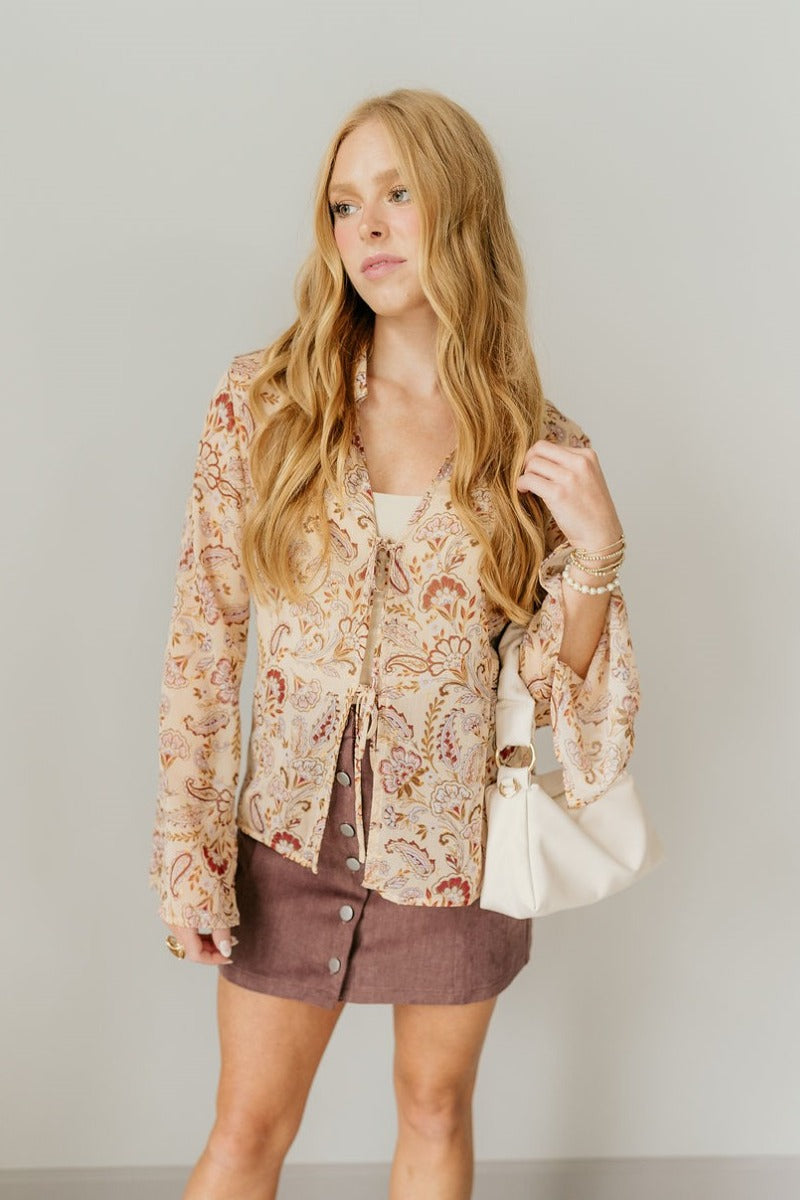 Front view of model wearing the Rosalee Printed Tie-Front Long Sleeve Top that has sheer, a paisley and flroal pattern with gold and silver metallic details, front tie closures, a collared neck, and long flare sleeves with slits.