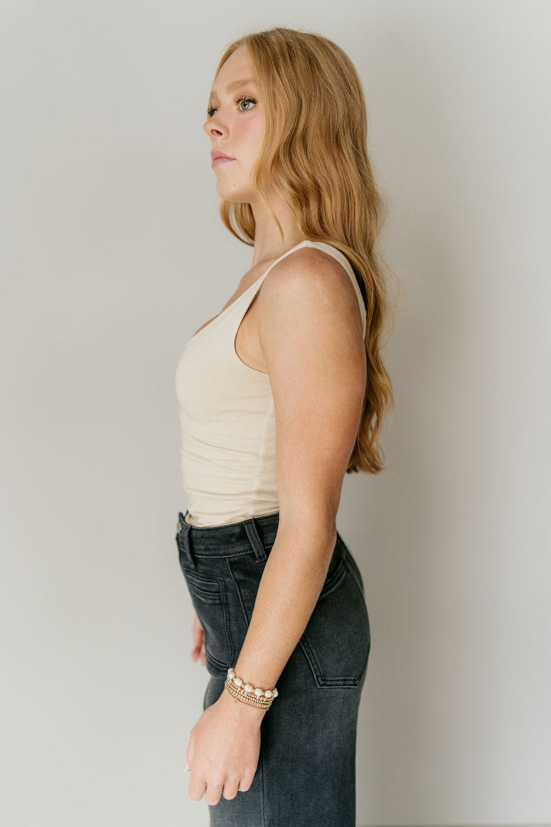 Side view of model wearing the Josie Beige Basic Scoop Neck Tank that has stretchy double-layered beige knit fabric, an elastic hem, a scooped neckline, and thick straps.